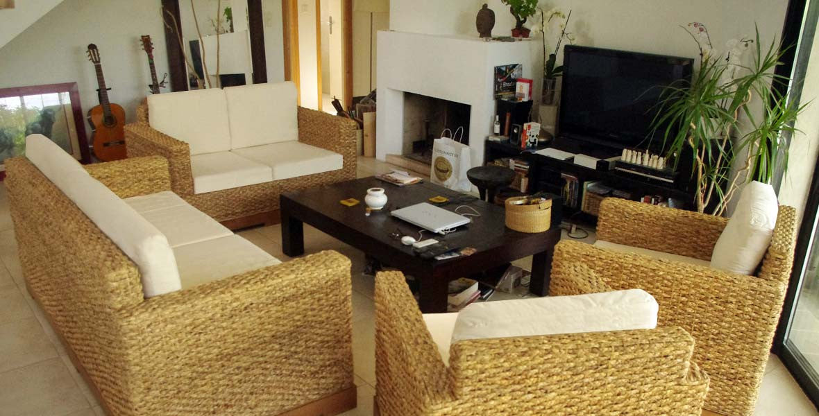 Salon accueillant avec canapé et fauteuils en rotin, table basse sombre et écran TV, ambiance naturelle et zen.